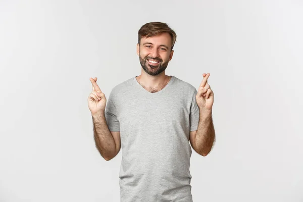 Retrato de hombre guapo optimista en camiseta gris, esperando algo con los dedos cruzados, pidiendo deseo y sonriendo, de pie sobre fondo blanco —  Fotos de Stock