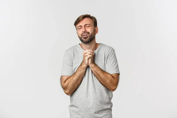 Retrato de homem bonito em camiseta cinza, implorando por algo, pleiteando e parecendo miserável, em pé sobre fundo branco — Fotografia de Stock