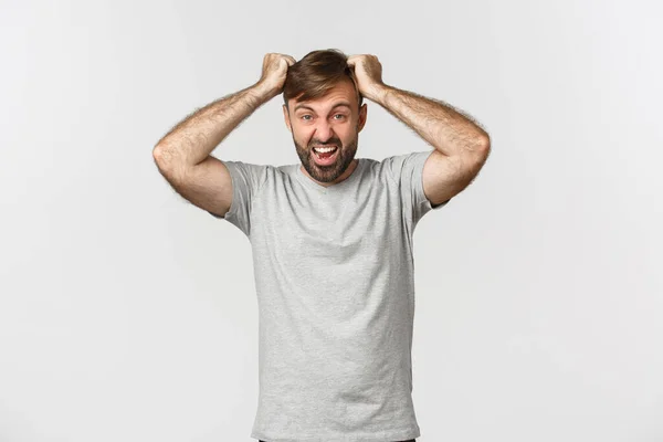 Porträtt av bekymrad galen kille i grå t-shirt, slita hår från ilska och skrika, står över vit bakgrund — Stockfoto