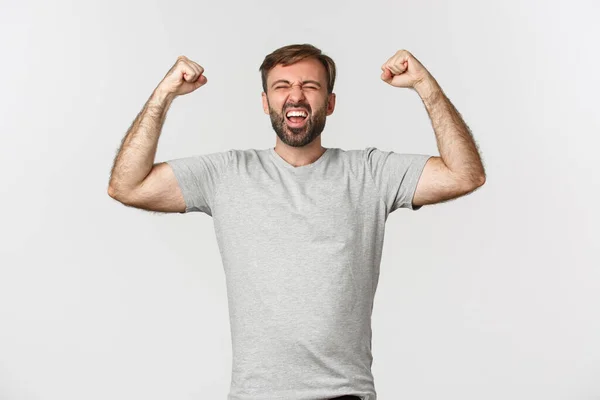 Portrait of happy guy feeling like a champion, raising hands up and rejoicing, shouting for joy, celebrating victory and triumphing, standing over white background — Stock Photo, Image