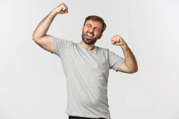 Muotokuva innoissaan ja onnellinen mies harmaa t-paita, jolloin nyrkki pumppu ja huutaa kyllä juhlii voittaa, voittoisa valkoinen tausta — kuvapankkivalokuva
