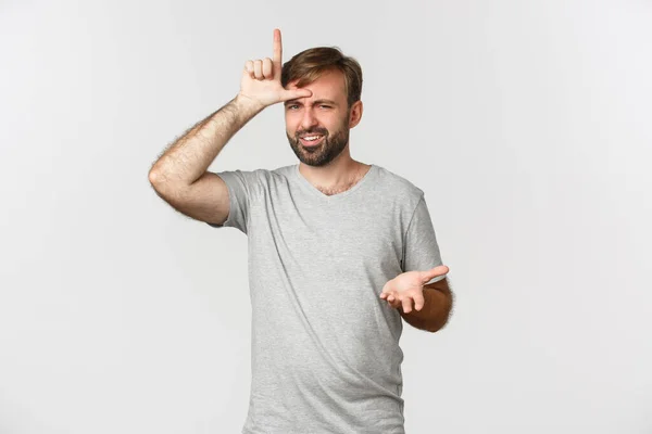 Retrato de homem arrogante em camiseta cinza, zombando de pessoa que perdeu, mostrando sinal perdedor na testa e olhando com desânimo, de pé sobre fundo branco — Fotografia de Stock