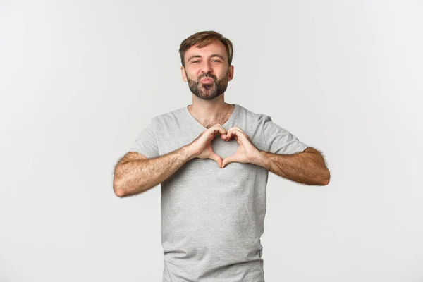 Retrato de belo homem sorridente com barba, mostrando sinal de coração, confessar no amor, beijando alguém, de pé sobre fundo branco — Fotografia de Stock