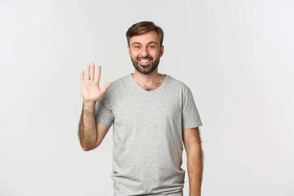 Портрет красивого кавказца в серой футболке, здоровающегося и приветливо машущего рукой, стоящего на белом фоне — стоковое фото