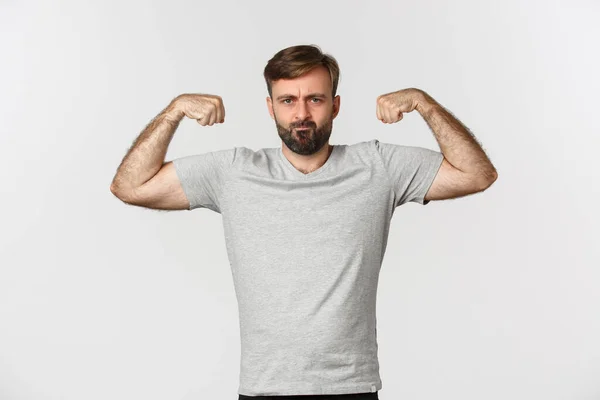 Muotokuva luottavainen parrakas mies taivutus hauislihas, osoittaa hänen lihaksia harjoittelun jälkeen, seisoo valkoisella taustalla — kuvapankkivalokuva