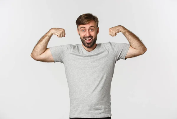 Портрет уверенного бородатого мужчины, сгибающего бицепсы, показывающего мышцы после тренировки в спортзале, стоящего на белом фоне — стоковое фото