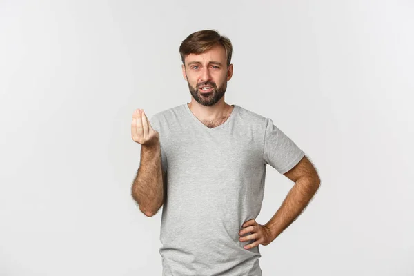 Молодой разочарованный мужчина что-то объясняет, трясет пальцами и смотрит в камеру, стоя в серой футболке на белом фоне — стоковое фото