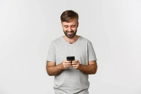 Porträtt av stilig modern kille med skägg, meddelanden och tittar på smartphone skärm, står över vit bakgrund — Stockfoto