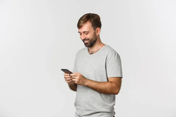 Porträtt av modern skäggig man i grå t-shirt, meddelanden och titta på smartphone glad, står över vit bakgrund — Stockfoto