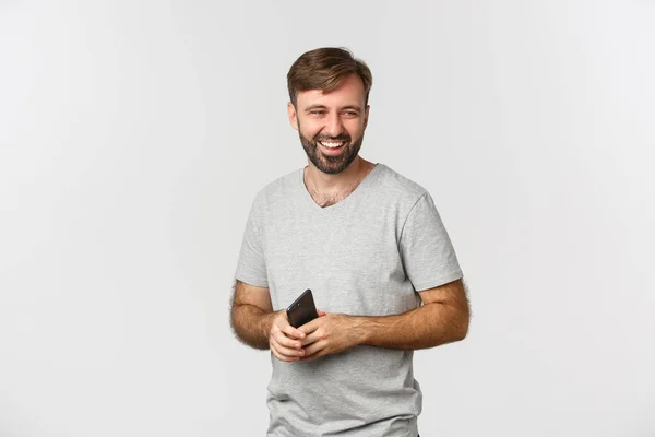 Изображение красивого бородатого мужчины в серой футболке, держащего смартфон, смотрящего направо и смеющегося, стоящего на белом фоне — стоковое фото