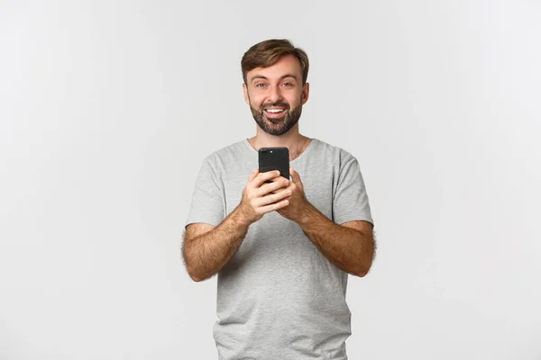 Χαμογελαστή τύπος με γενειάδα, λήψη φωτογραφιών ή εγγραφή βίντεο στο κινητό τηλέφωνο, στέκεται πάνω από το λευκό φόντο — Φωτογραφία Αρχείου