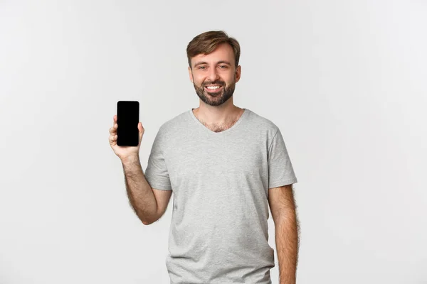 Portrét usmívajícího se pohledného muže v šedém tričku, zobrazující displej mobilního telefonu, doporučující aplikaci nebo nákupní stránku, stojící nad bílým pozadím — Stock fotografie