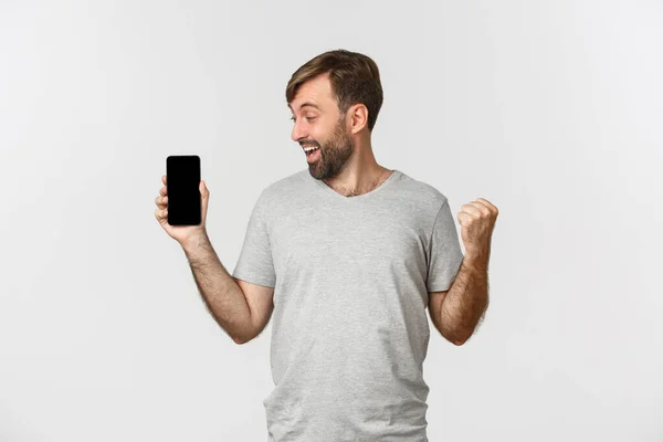 Portrét vzrušeného muže s vousy, dosažení cíle v aplikaci, zobrazení obrazovky mobilního telefonu a radosti, něco vyhrávat, stojící nad bílým pozadím — Stock fotografie