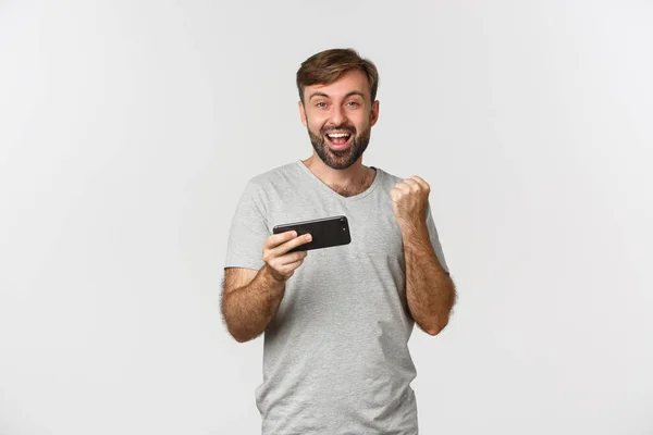 Porträtt av lycklig man i grå t-shirt, hålla smartphone och glädje, triumferande över prestation, står över vit bakgrund — Stockfoto