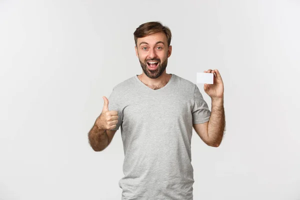 Portret van een knappe glimlachende man in grijs t-shirt, met kredietkaart, duimen omhoog in goedkeuring, over een witte achtergrond — Stockfoto
