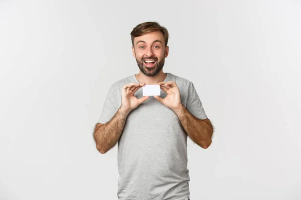 Iloinen valkoihoinen mies harmaassa t-paidassa, joka näyttää luottokorttia ja hymyilee, innoissaan ostoksille, seisoo valkoisen taustan yläpuolella — kuvapankkivalokuva