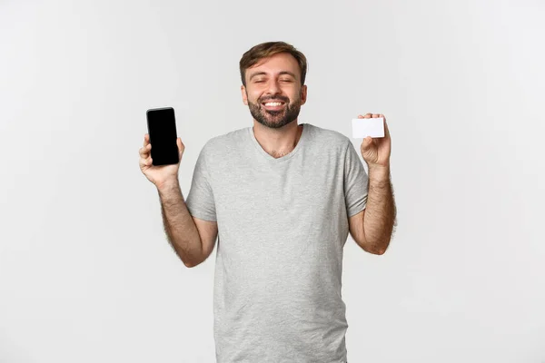 Fešák mladý muž nakupování on-line, držící kreditní kartu a mobilní telefon, ukazuje displej smartphone, stojící nad bílým pozadím — Stock fotografie