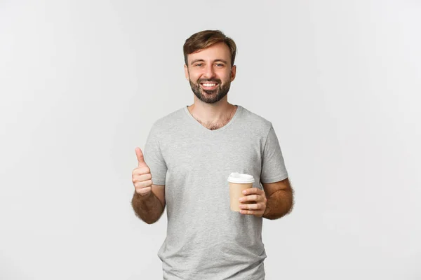 Portrait of cheerful caucasian man in gray t-shirt, drinking coffee and showing thumbs-up, like the drink, standing over white background — Stock Photo, Image