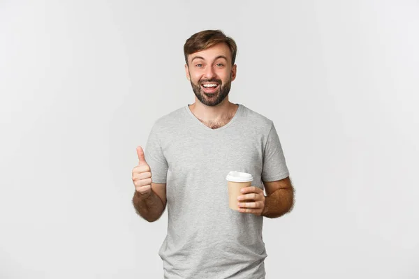 Retrato de homem caucasiano alegre em t-shirt cinza, beber café e mostrando polegar para cima, recomendar e como café, de pé sobre fundo branco — Fotografia de Stock