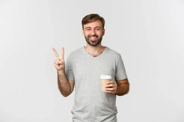 Imagem de cara feliz bebendo café takeaway, mostrando sinal de paz e sorrindo, de pé sobre fundo branco — Fotografia de Stock