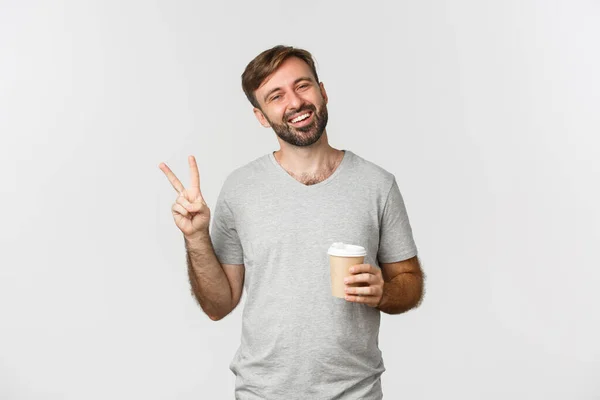 Portret van een zorgeloze glimlachende man in grijs t-shirt, koffie drinken en vredesbord, over een witte achtergrond staan — Stockfoto