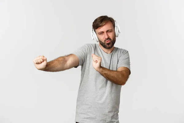 Ritratto di ragazzo bello felice in t-shirt grigia, ascoltare musica in cuffie senza fili e ballare, in piedi su sfondo bianco — Foto Stock
