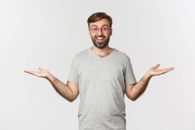 Sakallı, gri tişörtlü ve gözlüklü yakışıklı adam, ellerini yana açmış ve iki şey tutuyor, ürünleri gösteriyor, beyaz arka planda duruyor.