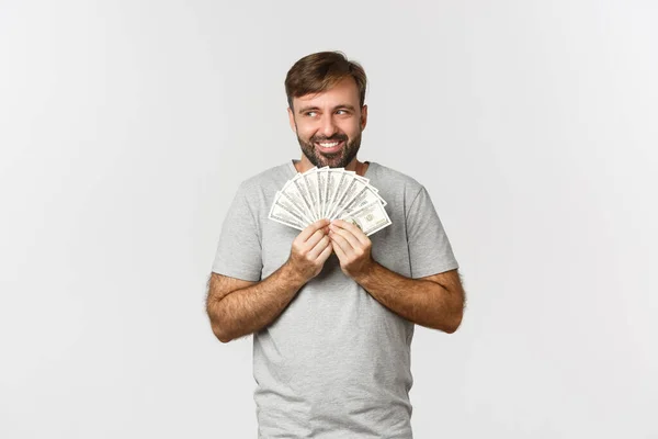 Chciwy uśmiechnięty chciwy mężczyzna z brodą, myślący o zakupach, trzymający pieniądze i patrzący w lewy górny róg, stojący nad białym tłem — Zdjęcie stockowe