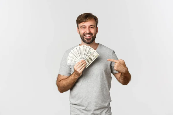 Retrato de guapo barbudo modelo masculino en camiseta gris, apuntando al dinero y sonriendo, consiguiendo un préstamo, de pie sobre fondo blanco — Foto de Stock
