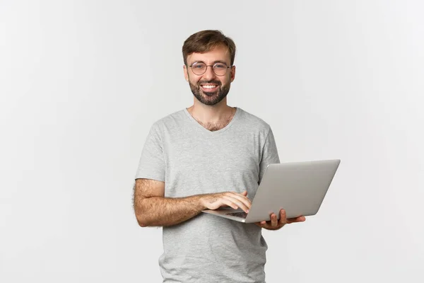 Jonge knappe freelancer in bril en grijs t-shirt, lachend blij, werken met laptop, staande over witte achtergrond — Stockfoto