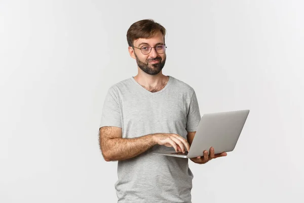Jonge zelfverzekerde freelancer in bril en grijs t-shirt, werken met laptop, glimlachen blij, staan over witte achtergrond — Stockfoto