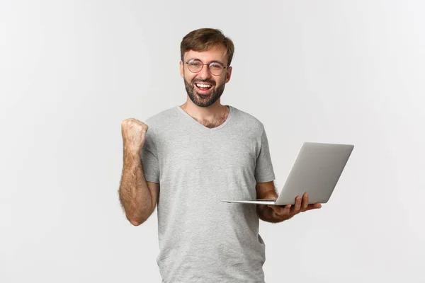 Retrato de cara bonito alegre em óculos e camiseta cinza, segurando laptop, alcançar objetivo, ganhar algo, de pé sobre fundo branco — Fotografia de Stock