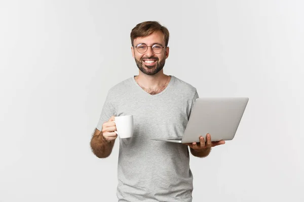 Jong gelukkig man werken met laptop, het drinken van koffie en glimlachen, staan over witte achtergrond — Stockfoto