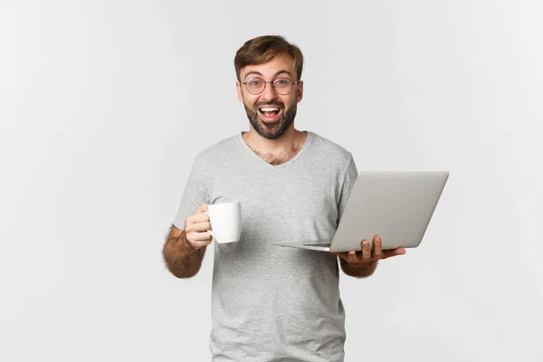 Jong gelukkig man werken met laptop, het drinken van koffie en glimlachen, staan over witte achtergrond — Stockfoto