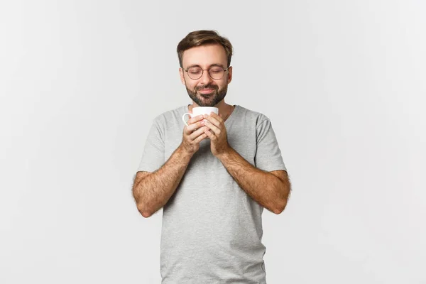 Porträtt av glad leende man i glasögon och avslappnad t-shirt, blunda och lukta kaffe i mugg, står över vit bakgrund — Stockfoto