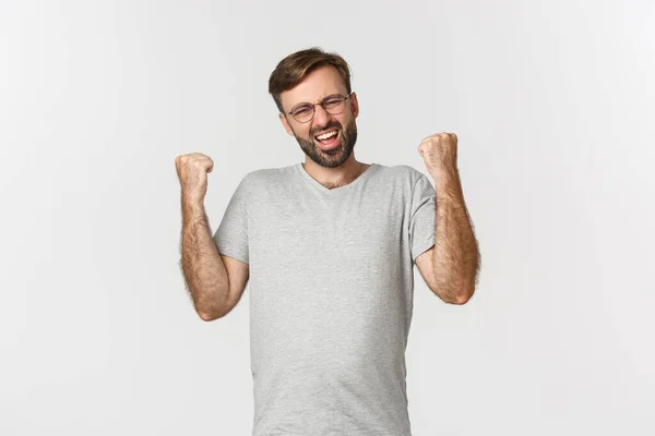 Bild av upphetsad man i grå t-shirt och glasögon, vinna något, glädje och känsla som mästare, står över vit bakgrund — Stockfoto