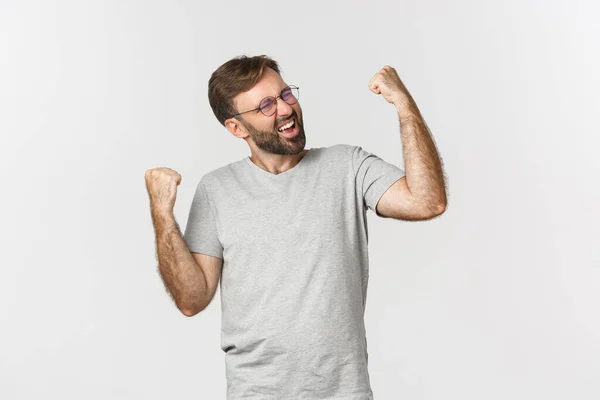 Imagen del hombre excitado en camiseta gris y gafas, ganando algo, regocijándose y sintiéndose como campeón, de pie sobre fondo blanco —  Fotos de Stock