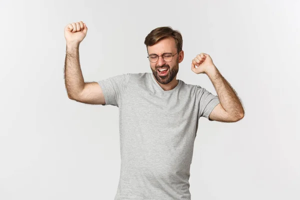 Imagen del hombre excitado en camiseta gris y gafas, ganando algo, regocijándose y sintiéndose como campeón, de pie sobre fondo blanco — Foto de Stock