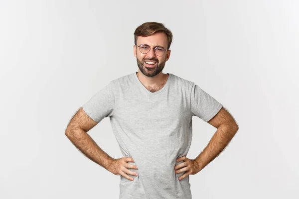 Image of confused bearded man in glasses and gray t-shirt, looking at something strange, frowning perplexed, standing over white background — Stock Photo, Image