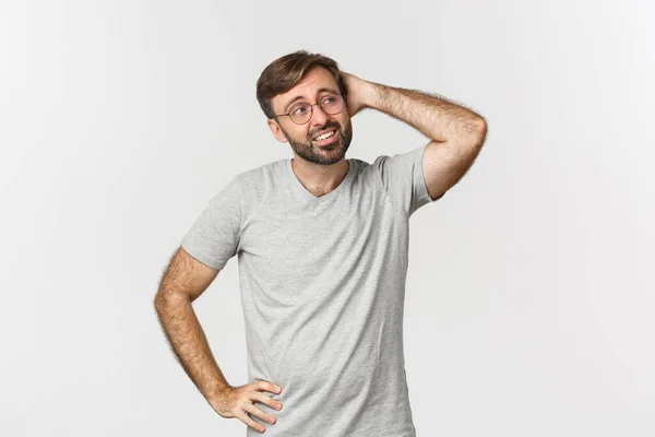 Porträtt av obeslutsam vuxen man tänker, klia huvudet och ser förvirrad på övre högra hörnet, står i grå t-shirt och glasögon över vit bakgrund — Stockfoto