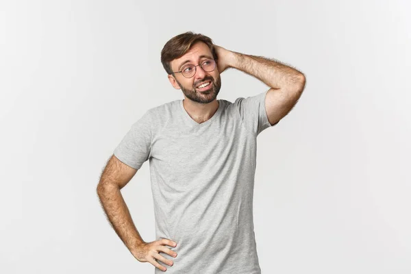 Retrato de homem adulto indeciso pensando, coçando a cabeça e olhando confuso no canto superior direito, de pé em camiseta cinza e óculos sobre fundo branco — Fotografia de Stock