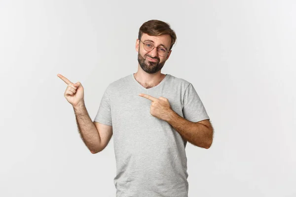 Skeptinen ja huvittamaton parrakas mies virnuilee, osoittaa sormella vasempaan yläkulmaan, näyttää logoa, seisoo valkoisen taustan päällä — kuvapankkivalokuva
