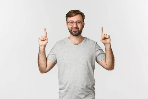 Homem barbudo alegre sorrindo, apontando os dedos para cima, mostrando logotipo, vestindo camiseta cinza, de pé sobre fundo branco — Fotografia de Stock