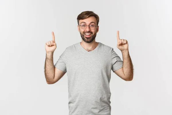 Homem barbudo alegre sorrindo, apontando os dedos para cima, mostrando logotipo, vestindo camiseta cinza, de pé sobre fundo branco — Fotografia de Stock