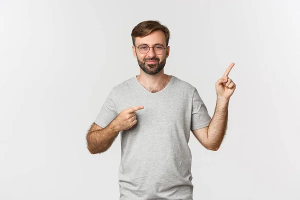 Cheerful bearded man smiling, pointing fingers at upper right corner, showing logo, standing over white background — Stock Photo, Image