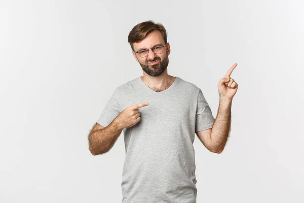 Disappointed bearded man grimacing, pointing fingers at upper right corner, showing logo, standing over white background — Stock Photo, Image