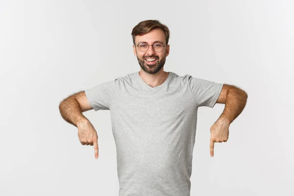 Homem barbudo alegre sorrindo, apontando os dedos para baixo, mostrando logotipo, vestindo camiseta cinza, vestindo camiseta cinza, de pé sobre fundo branco — Fotografia de Stock