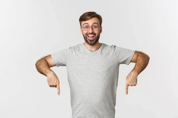 Hombre barbudo alegre sonriendo, señalando con los dedos hacia abajo, mostrando el logotipo, usando camiseta gris, usando camiseta gris, de pie sobre fondo blanco — Foto de Stock