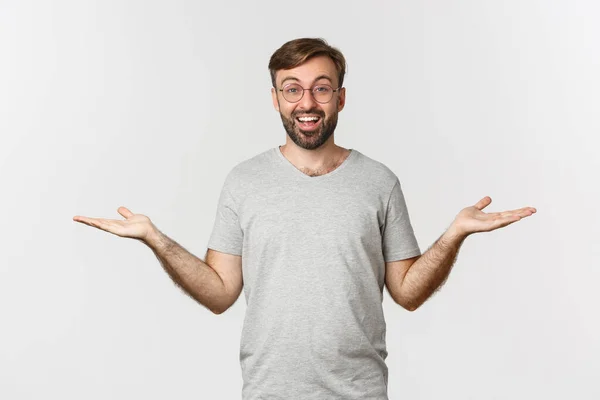 Handsome man with beard, wearing gray t-shirt and glasses, spread hands sideways and holding two things, demonstrate products, standing over white background — Stock Photo, Image