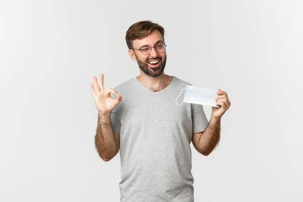 Conceito de pandemia, coronavírus e distanciamento social. Imagem de cara bonito sorrindo em óculos, mostrando sinal ok e recomendando usar máscara médica — Fotografia de Stock